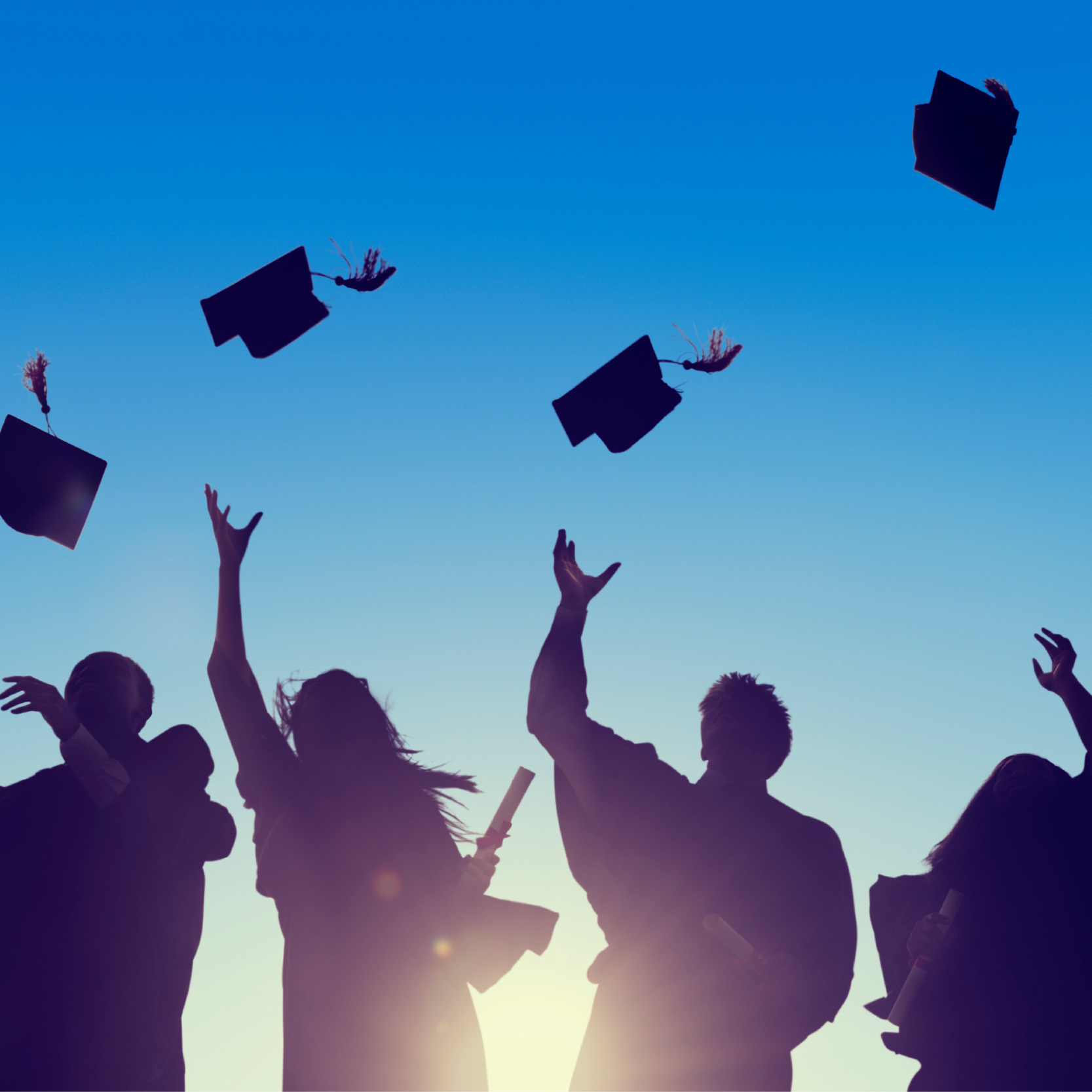 Graduates in silhouette from the sun throw their hats